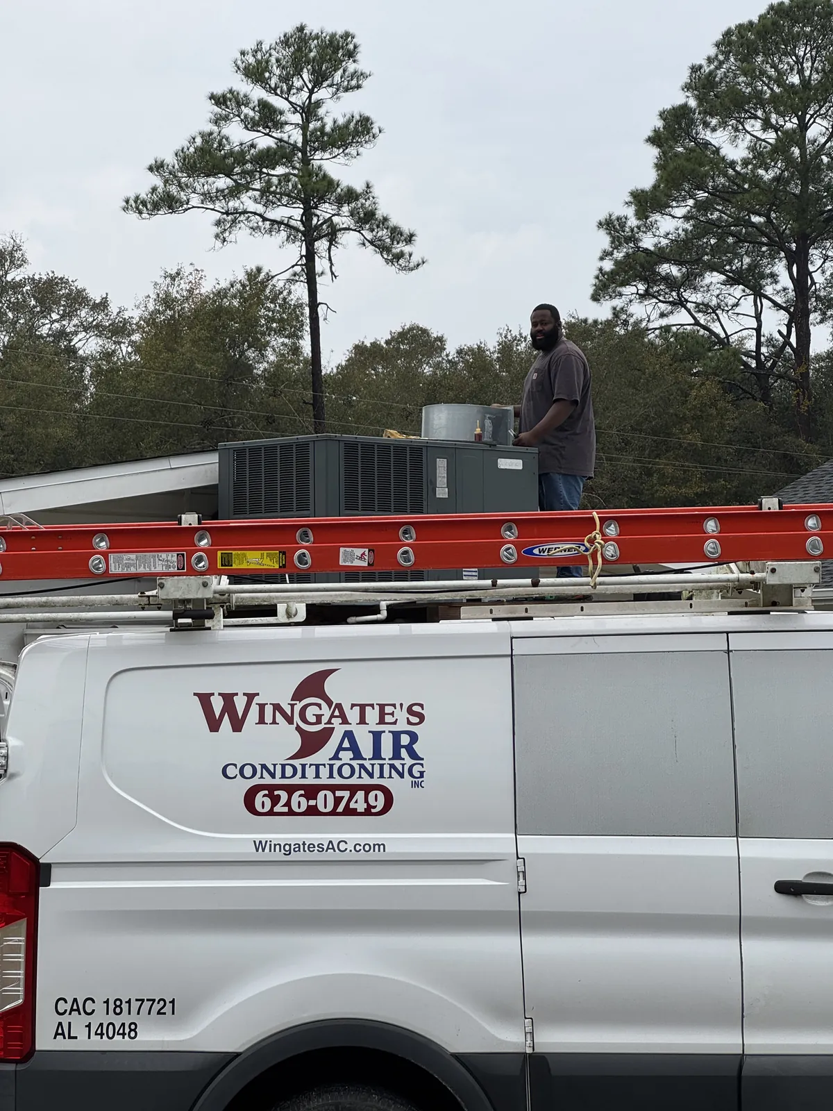A Wingate's Air Conditioning technician working on a rooftop package unit with the company van parked below